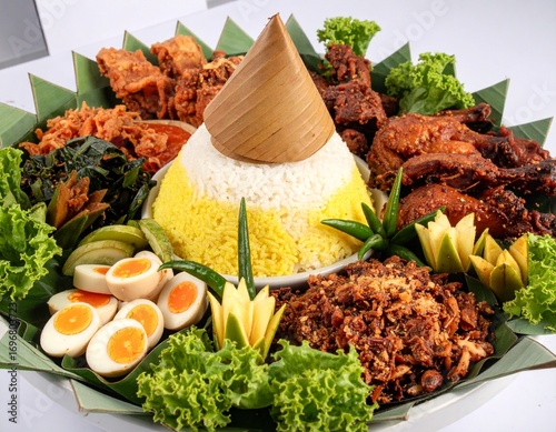 Traditional Indonesian Rice Tumpeng with many topping like egg, vegetables, tempeh, chicken, chili, sambal, cucumber, noddle, yellow rice and make mountain shape in the middle with banana leaf as base