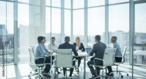 Blurred business meeting of professionals seated around a conference table in modern office with large glass windows overlooking the city.