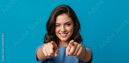 You can do this. Beautiful cheerful brunette girl winking and smiling, pointing fingers at camera, praising good job, inviting to event, standing against blue background