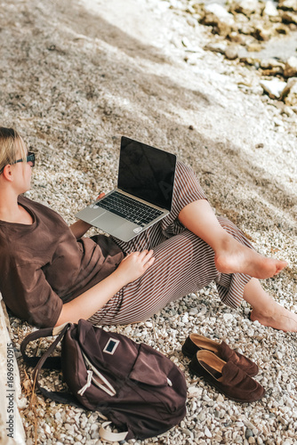 Young woman working remotely by the sea with a laptop and solar panel, embracing sustainable travel, mindful lifestyle and digital freedom. Concept of new work, balance and conscious living.