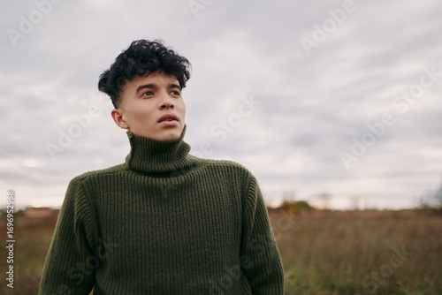 A young man in a green turtleneck stands in an open field, portrait captures an authentic, credible candid moment with natural expression and relaxed posture under cloudy sky.