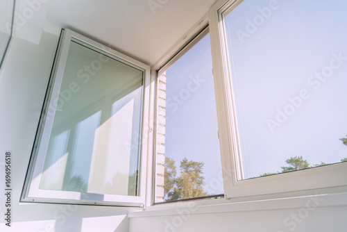 An open white plastic window in a bright, minimalist room, letting in sunlight and a summer breeze. The serene view from the apartment looks out on a clear blue sky and green trees.