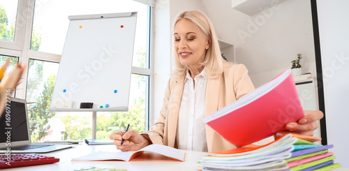 Mature female teacher with copybooks at table in classroom