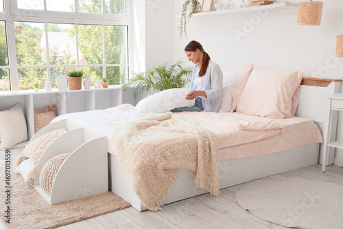 Young woman arranging pillows on a modern bed with pink bedding in a bright, ...