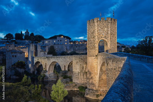Foto Fortified, historic Pont de Besalú Bridge over the Fluvià River illuminated at blue hour in Catalonia, Spain, with moody sky and stone arches