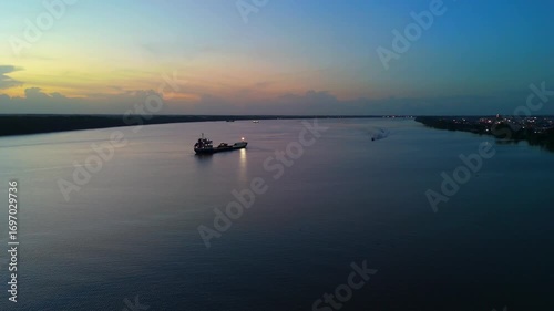 Aerial view of a wide river at dusk with a large cargo ship in the middle creating a beautiful atmosphere. 4K 60fps