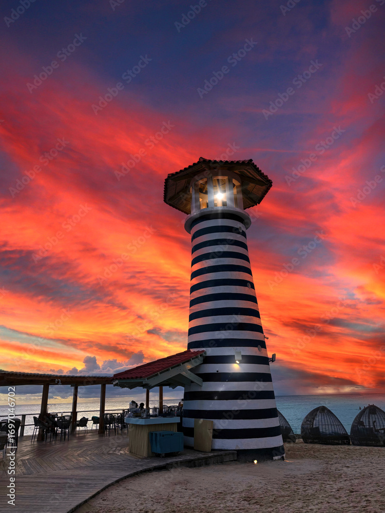 Obraz premium Striped lighthouse illuminating the beach bar with intense red and orange sunset sky