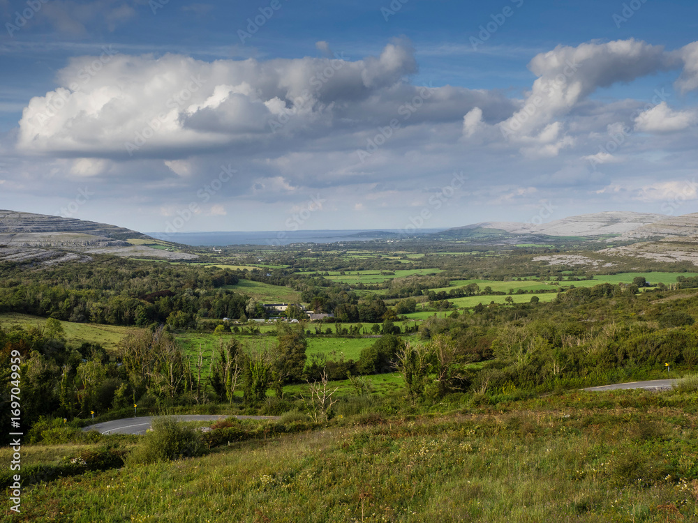 Obraz premium Landscape scene with green fields, hills, ocean and blue sky. Burren area, county Galway, Ireland. Warm sunny day. Rural Irish scenery in unique location. Travel and tourism.