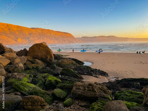 Sandy beach at sunset, with large rocks covered with green algae in foreground. Several surfers carrying their surfboards are walking along sandy shore, while hill in background glows in golden shade.