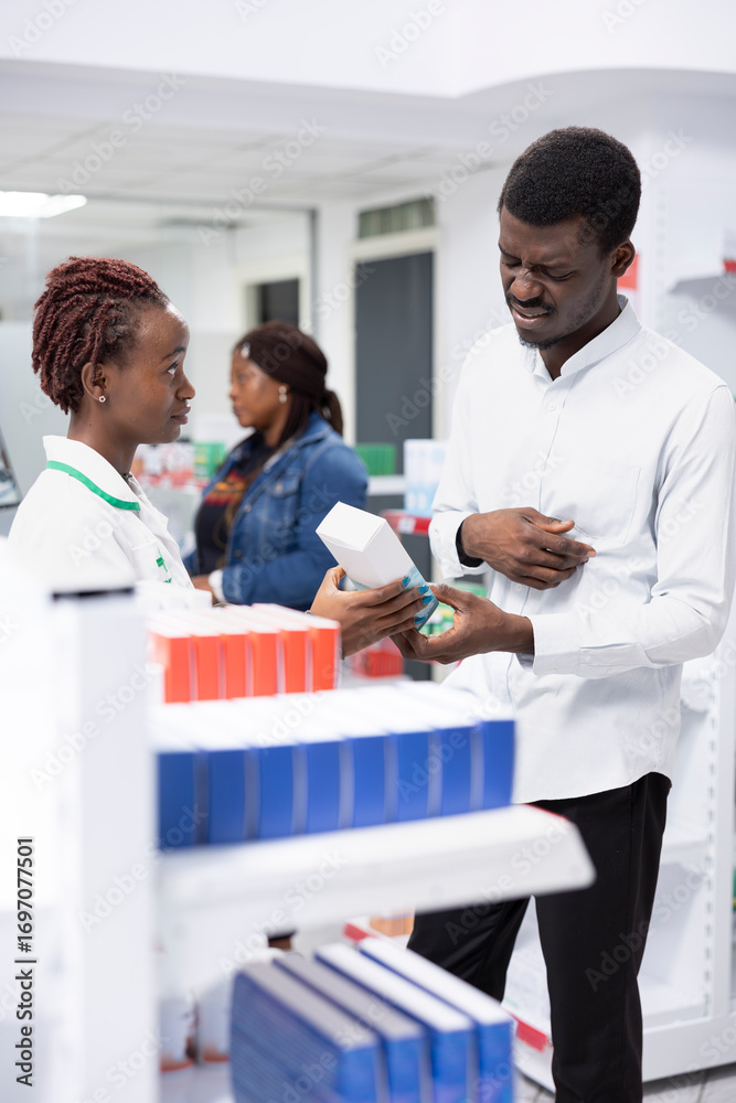 Fototapeta premium African american expert offers medication for a guy with abdominal pain, assisting client in treating a digestive inflammation or food poisoning. Pharmaceutical care in a retail setting.