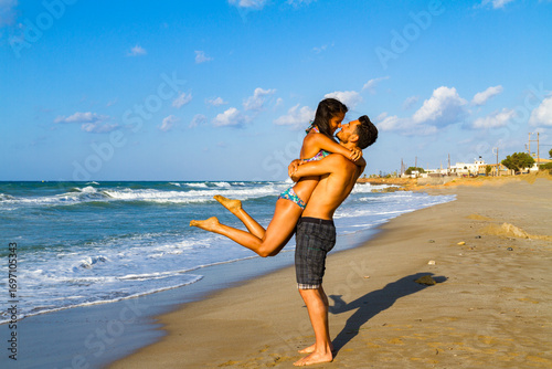Happy young couple in bikini and shorts enjoying summer at the beach at dusk, breathing fresh air, kissing and teasing one another