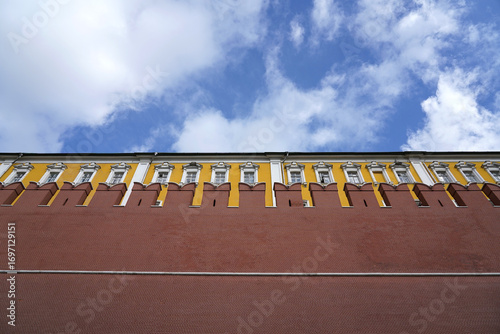 A red old vintage antique brick wall with battlements and a yellow antique building against a blue sky with clouds   