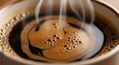 Close-up of a steaming cup of coffee in the foreground. The coffee has a dark, rich color with a frothy surface and visible bubbles, indicating freshness.