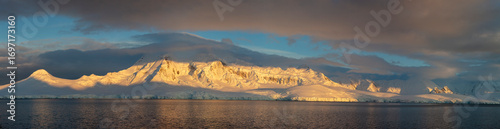 Panoramic view of Antarctic mountains bathed in golden light during sunrise or sunset, with dark clouds and calm ocean waters creating a dramatic contrast. A breathtaking scene of the polar wilderness