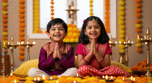 Two children dressed in colorful ethnic wear sit indoors surrounded by marigold flowers and diyas, performing a prayer during Diwali celebrations.