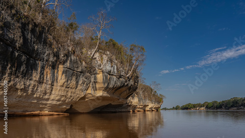 A calm red-brown African river. Trees grow on the steep limestone eroded shores. Blue sky, clouds. Reflection on the surface of the water. Madagascar. Manambolo river.