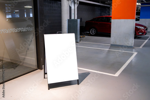 Blank white mockup signboard placed in an indoor parking garage, with cars parked in the background. Empty board with copy space for advertising, signage, branding, and urban infrastructure.