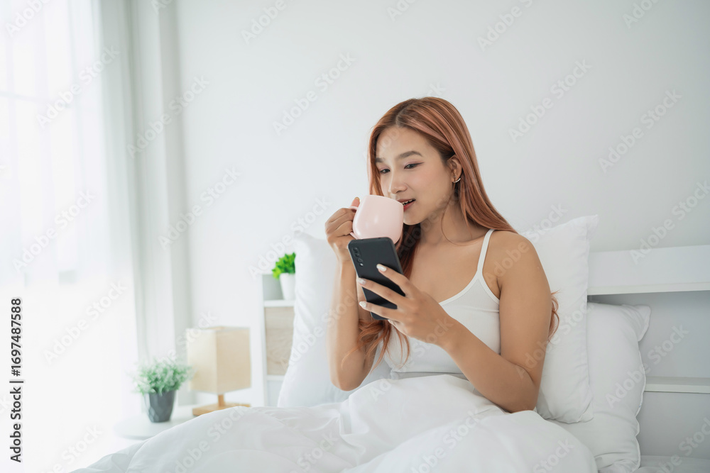 © WMSTUDIO - Young woman enjoying morning beverage in bed while using smartphone for browsing or social media in bright sunlit room with modern decor