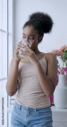 african american teenage girl looking outside window while holding glass of water in peaceful moment expressing beauty glow wellness and calm lifestyle through hydration and natural indoor light