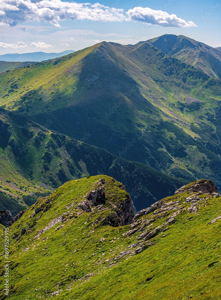 Fototapeta premium Scenic mountain ridge with green grassy slopes, rocky outcrops, and distant peaks under a blue sky. Natural alpine landscape ideal for hiking and nature lovers.