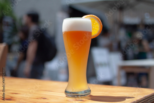 A freshly poured wheat beer served in a tall glass, garnished with a vibrant orange slice, and set on a rustic wooden table in a sunny outdoor setting.