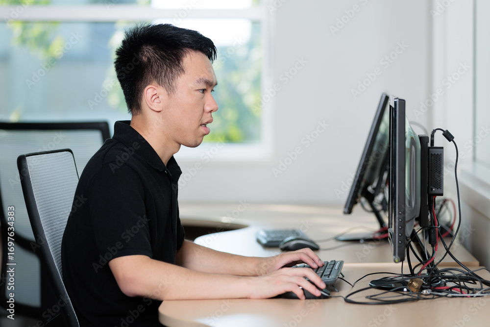 Fototapeta premium Young man types on a keyboard while focused on a desktop monitor in a computer lab classroom. Side profile view shows concentration in task.
