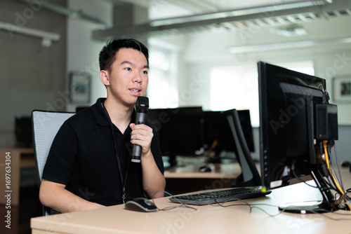 Fotomural A young man sits in front of a desktop computer in a classroom computer lab, holding a microphone while speaking, responding thoughtfully during a discussion or presentation