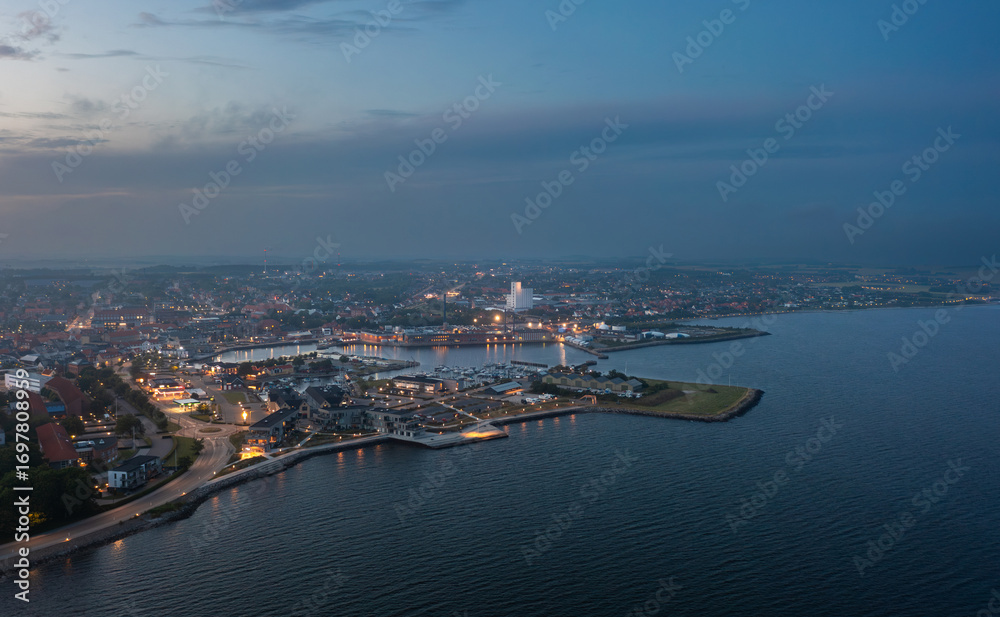 Obraz premium Aerial night view of Thisted, Denmark. Town lights, marina, Limfjord coastline, and surrounding neighborhoods create a tranquil Nordic cityscape under a twilight sky