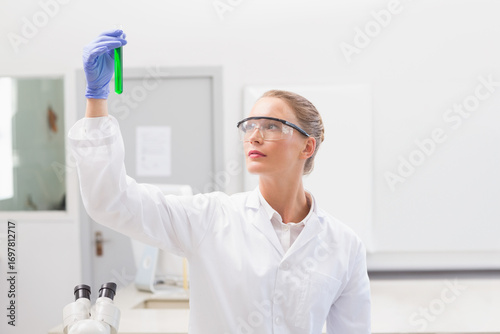 Scientist wearing lab coat, goggles and gloves holding green liquid test tube by microscope in lab
