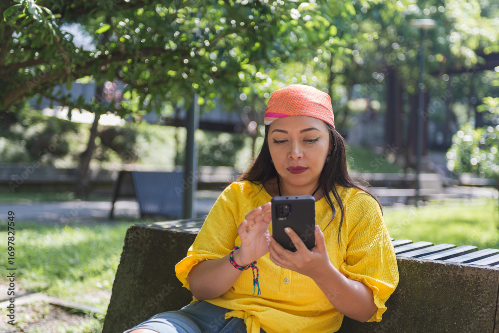 Fototapeta premium Woman in a yellow shirt and orange headscarf sits on a park bench, attentively using her smartphone