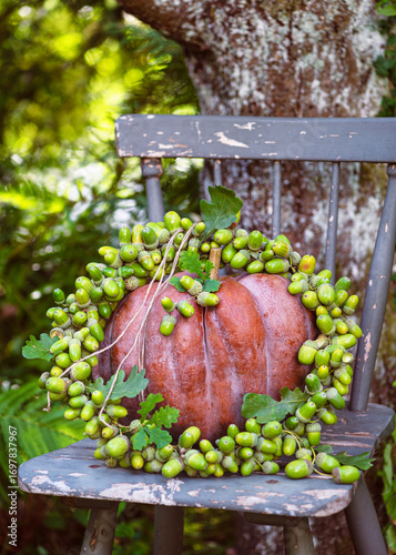 Rustic autumn still life with a deep orange pumpkin crowned by handmade vibrant wreath of green acorns, resting on a weathered chair beneath a tree. Vintage floral arrangement or garten decoration