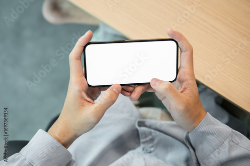 Blank screen smartphone holding horizontally with both hand while sitting at wooden table in a cafe.