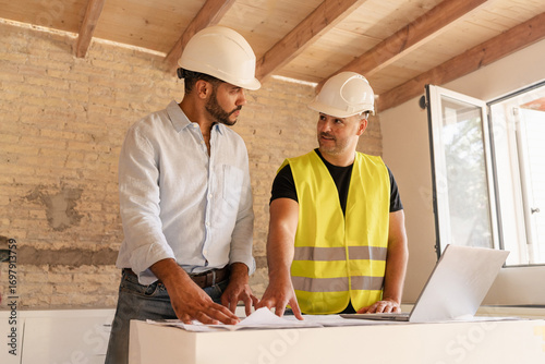 Male Contractor and engineer discussing blueprint using laptop during building construction