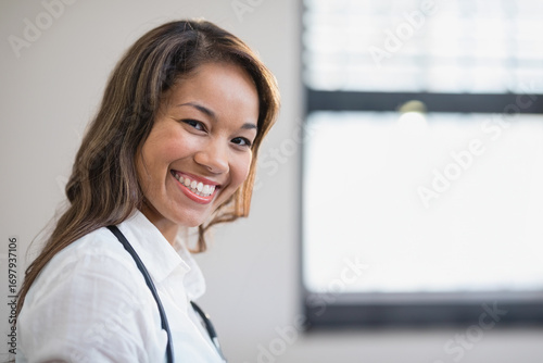 Mid adult Asian doctor wearing stethoscope standing in medical office smiling at camera, copy space Free Download Mid adult Asian doctor wearing stethoscope standing in medical office smiling at camera, copy space - GFXLoad