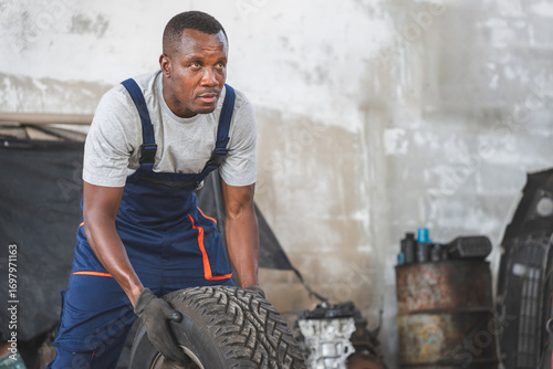 Foto Auto Mechanic Lifting Tire in a Garage, Tired Mechanic Working in an Automotive
