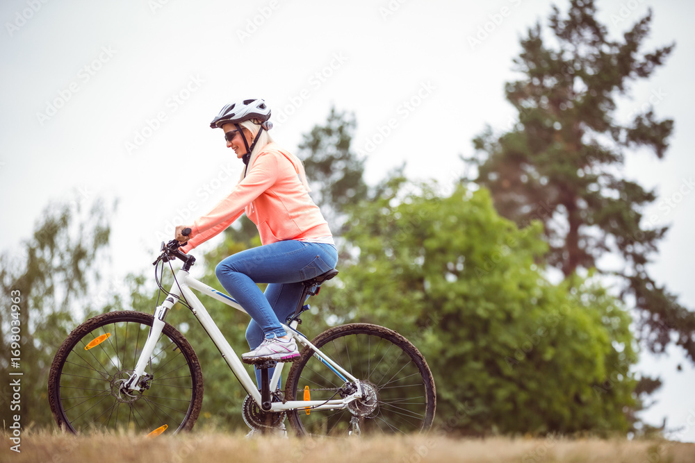 Naklejka premium Woman riding white mountain bicycle up grassy hill at forest edge wearing helmet and sunglasses