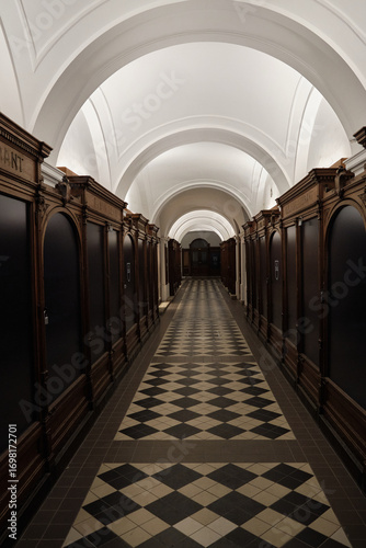 Arched corridor in a historic Viennese building featuring carved wooden shelves, black-and-white diamond tile floor, and ornate white arched ceiling in a grand interior.