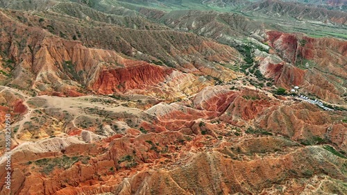 Drone flies forward above Skazka Canyon in Kyrgyzstan, revealing a breathtaking landscape of vivid red mountains and dramatic terrain in warm sunlight.