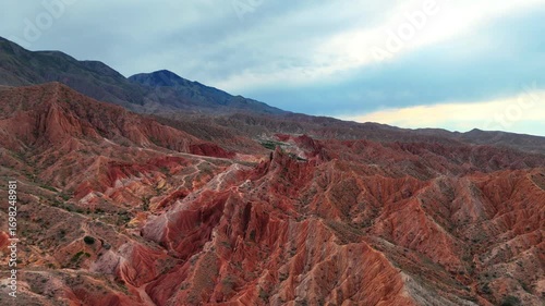 Drone video over sharp red peaks of Skazka Canyon in Kyrgyzstan, filmed on a cloudy day. The landscape features dramatic rock formations and rich earthy colors.