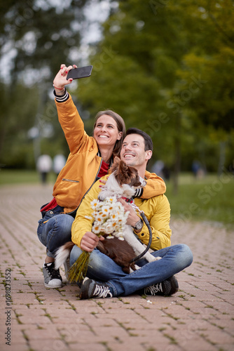 caucasian woman taking selfie with her boyfriend and his dog in a lap, sitting on a path in park
