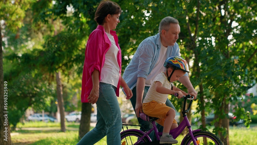 Fototapeta premium Child cyclist with parents in city park. Family weekend, father teaches son to ride bike in park. Happy family, little child in helmet with father and mother learning to ride bike outdoors in summer