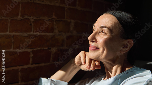 Fotografi Mature woman in white shirt smiling towards sunlight near brick wall