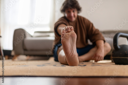 Man stretching foot with kettlebell, home workout, modern room
