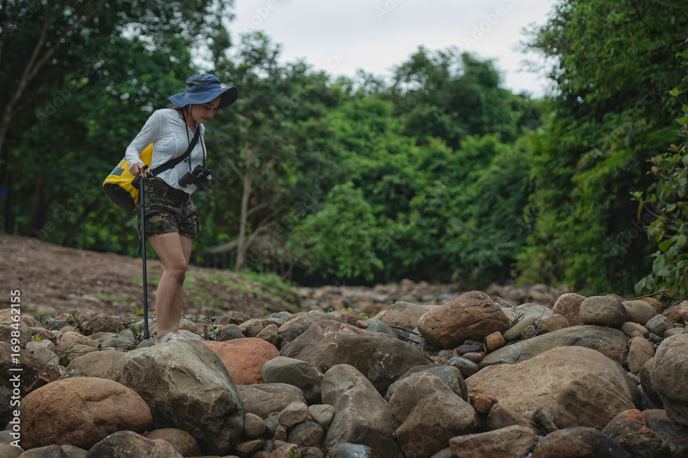 Obraz premium Young woman carrying a yellow backpack and camera is hiking through nature, carefully crossing a rocky stream in the forest. Outdoor adventure and eco-travel lifestyle concept. 