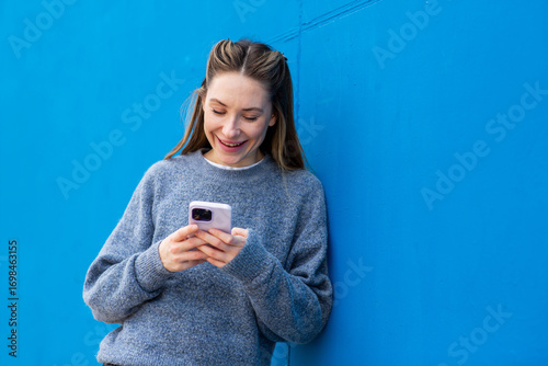 Smiling young woman leaning against blue wall and using mobile phone