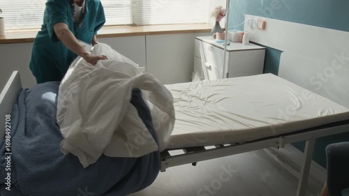 Zoom in shot of young nurse wearing scrubs removing pillow, sheet and blankets off patients bed after their discharge from hospital