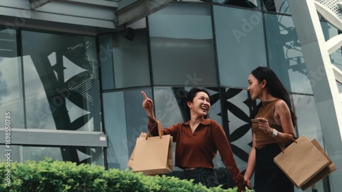 Two young women holding hands and shopping bags, walking and happily pointing at something near a modern mall