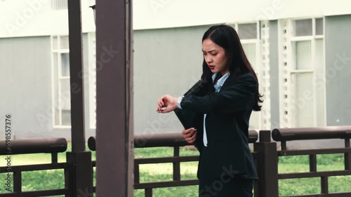 Professional businesswoman hurrying through sleek corporate lobby, glancing anxiously at wristwatch, signaling time-critical professional pressure