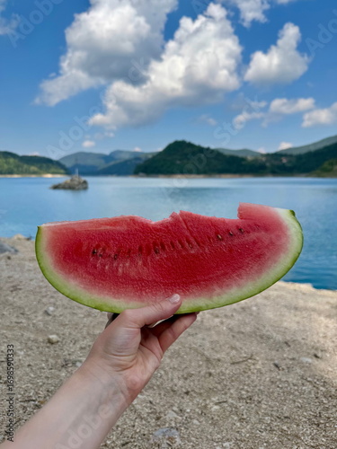 Close-up of a woman's hand holding a slice of watermelon with a beach and water in the background, distant mountains visible, summer outdoor scene, vacation and refreshing vibe