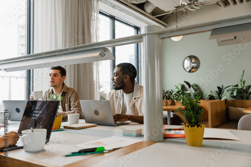 Two male workers sit at desks in front of laptops while one of them smiles and the other holds a cup